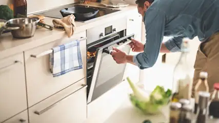 Ein Mann öffnet einen Ofen in einer hellen Küche. Auf der Theke stehen Kochutensilien und Zutaten.