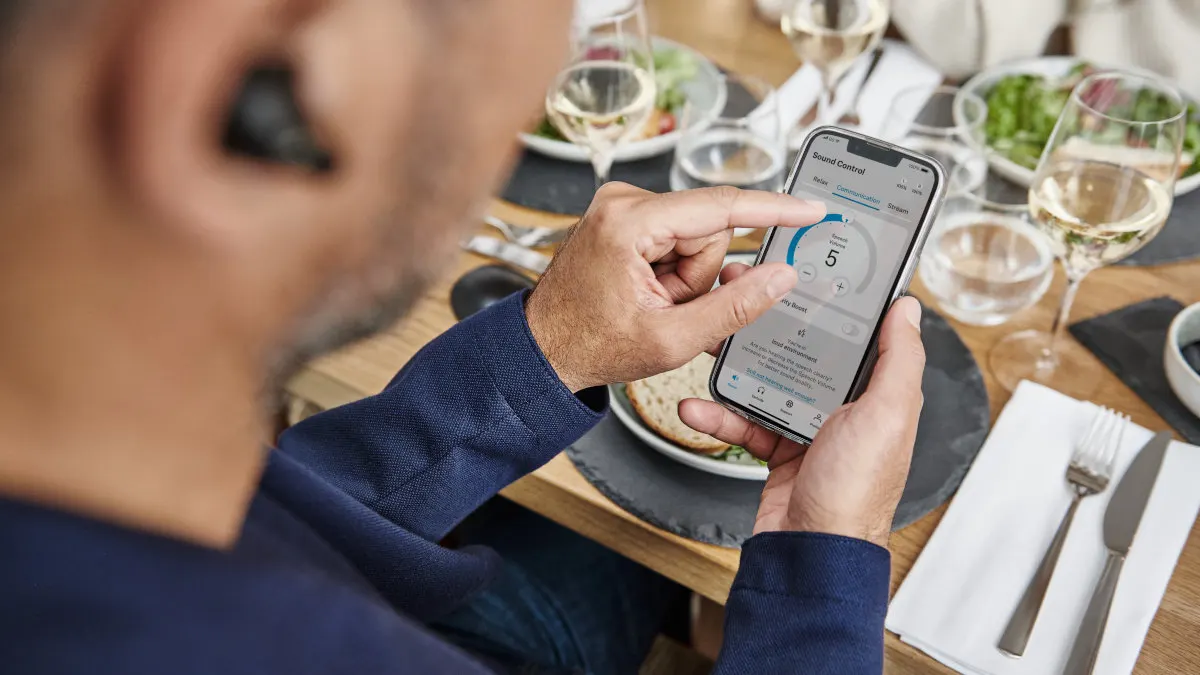 Ein Mann mit Hörgerät bedient eine App auf einem Smartphone am Tisch, umgeben von Speisen und Getränken.