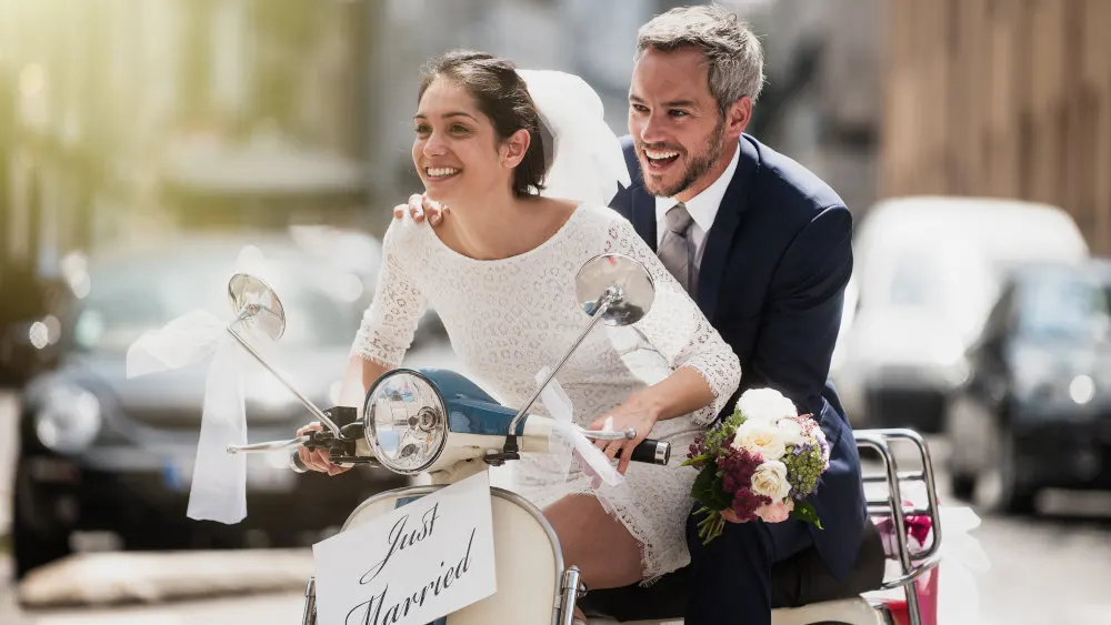 Ein Paar fährt auf einem hellblauen Roller mit "Just Married"-Schild. Die Frau trägt ein weißes Kleid, der Mann einen Anzug.