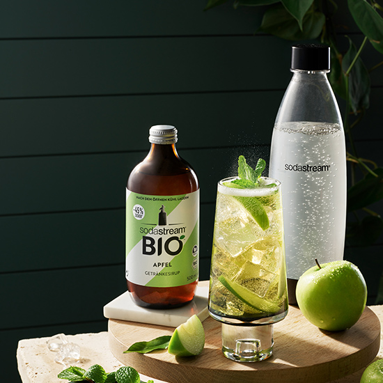 Eine Flasche Apfelsirup, ein Glas Apfelgetränk, eine Sodawasserflasche und ein grüner Apfel stehen auf einem Tisch.