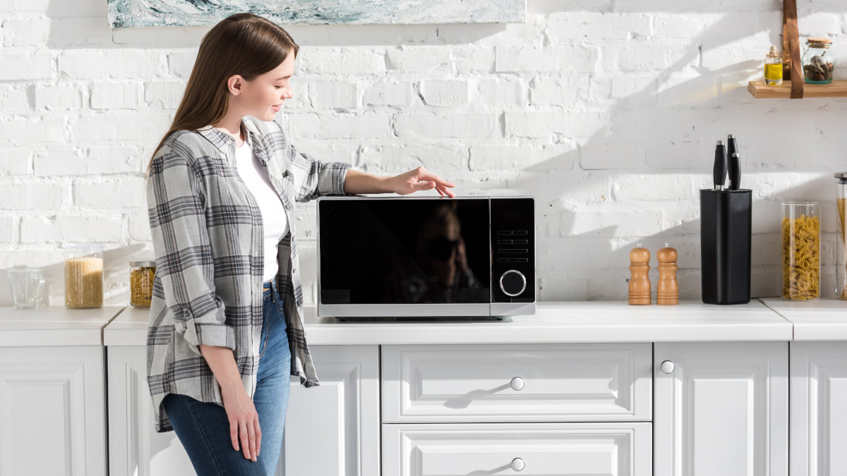 Eine junge Frau steht in einer hellen Küche und legt ihren Arm auf eine moderne Mikrowelle.