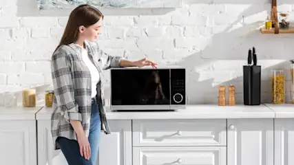 Eine junge Frau steht in einer hellen Küche und legt ihren Arm auf eine moderne Mikrowelle.