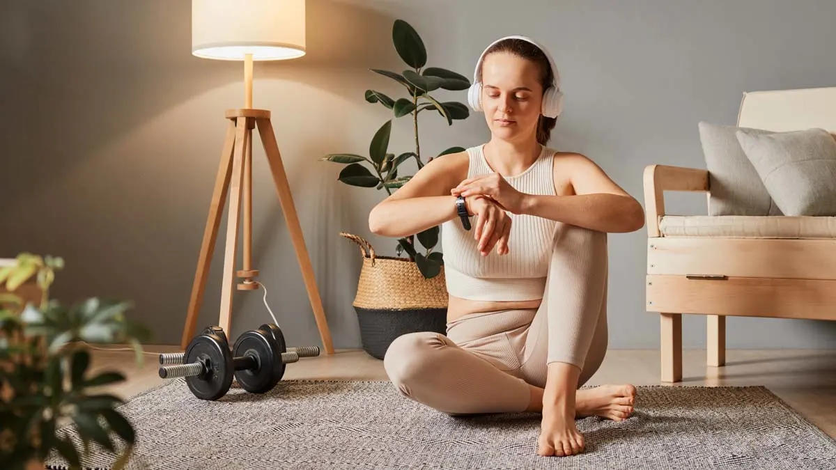 Eine Frau in Sportkleidung sitzt neben Kurzhanteln im Wohnzimmer und schaut auf ihren Fitnesstracker.