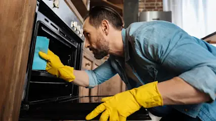 Ein Mann mit gelben Handschuhen wischt einen schwarzen Ofen mit einem blauen Tuch in einer Küche ab.