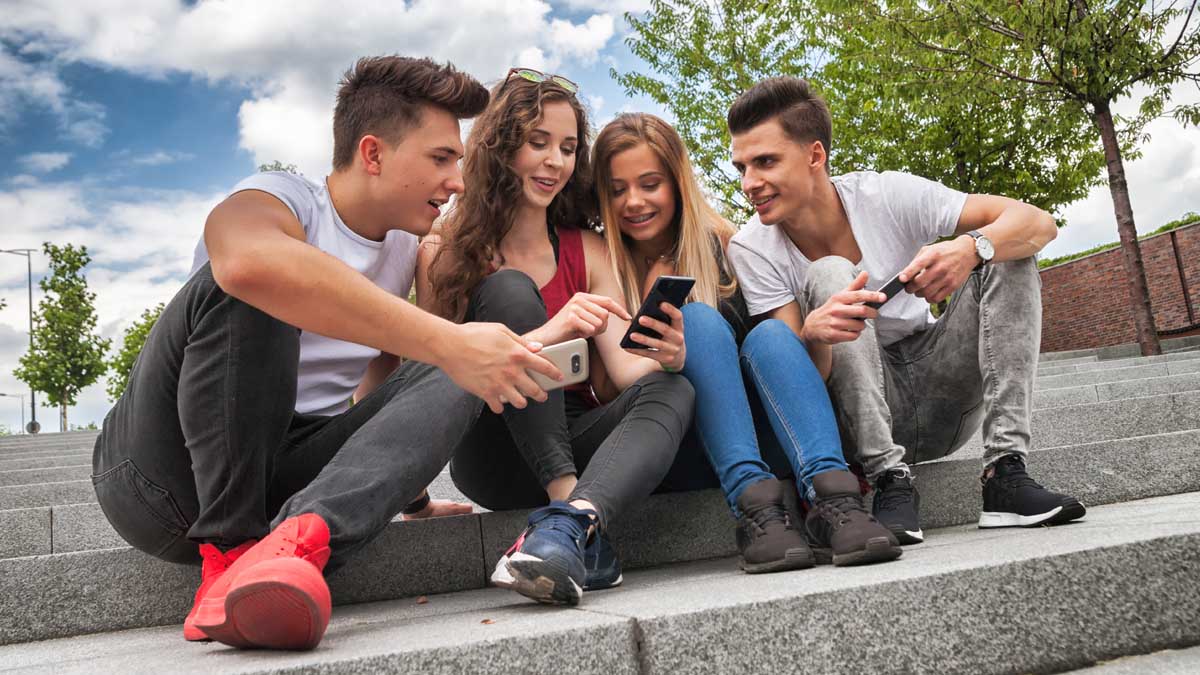 Eine Gruppe aus jungen Männern und Frauen sitzt mit ihren Smartphones in der Hand draußen auf einer Steintreppe. Sie sehen alle auf eines in der Mitte.
