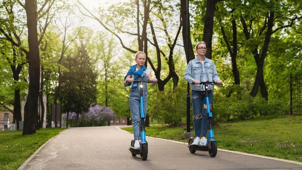 Mutter und Kind fahren auf E-Scootern einen Weg entlang. Im Hintergrund sind Bäume und Wiesen in einem Park sichtbar.