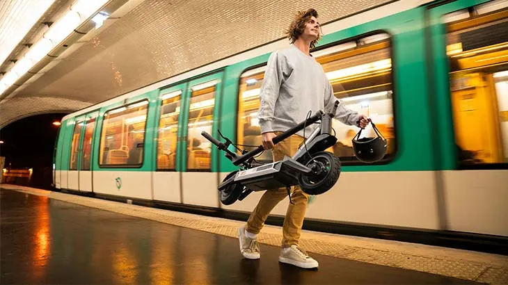 Ein Mann trägt einen grauen Roller und einen schwarzen Helm vor einem grünen und beigen Zug in einem Bahnhof.