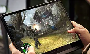 Person hält Tablet mit Videospielszene in Waldumgebung. Eine Spielfigur zielt mit einer Waffe, Explosionen sind sichtbar.