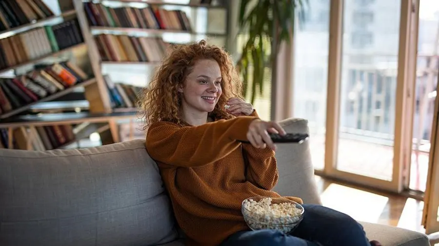 Eine Frau mit braunem Pullover sitzt mit Popcorn auf einem grauen Sofa und hält eine Fernbedienung in der Hand. Im Hintergrund Bücherregal und Fenster.