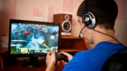 Ein Mann mit Headset spielt ein Videospiel auf einem Monitor mit einem Controller in seinen Händen. Lautsprecher sind im Hintergrund.