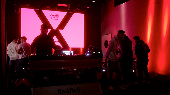 In einem dunklen Raum stehen Personen vor einem DJ-Pult mit roter Neon-Beleuchtung. Ein großes, rosa Logo ist im Hintergrund sichtbar.