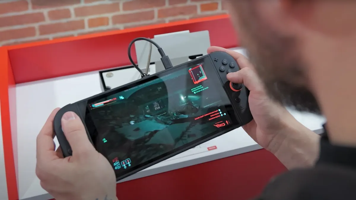 Eine Person hält ein schwarzes Handheld-Spielgerät mit einem laufenden Videospiel in der Hand. Das Gerät ist an ein Kabel angeschlossen.