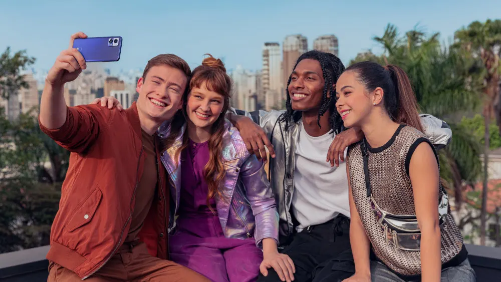 Eine Gruppe von vier Jugendlichen, die sich umarmen und mit einem blauen Mobiltelefon ein Selfie vor einer Stadtkulisse machen.