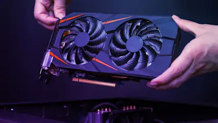 Eine Grafikkarte mit zwei Lüftern wird über einen Computergehäuse gehalten. Die Karte ist schwarz mit orangefarbenen Akzenten.