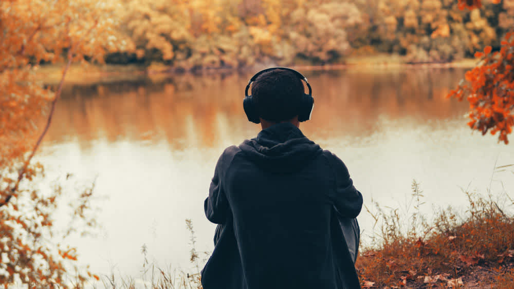 Mann sitzt vor einem See und hört Musik über Bluetooth-Over-Ear-Kopfhörer