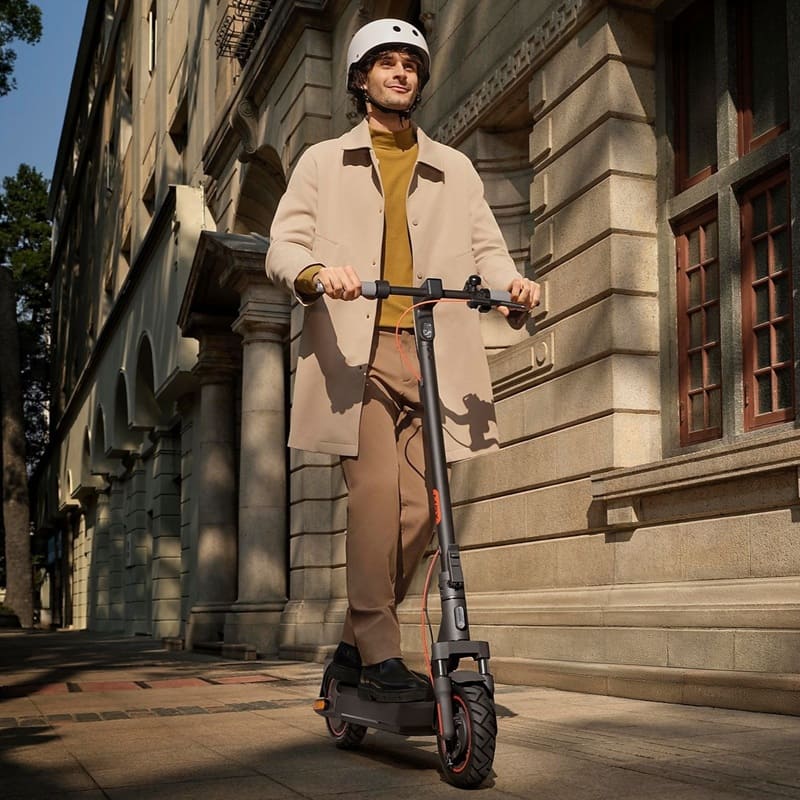 Ein Mann mit weißem Helm fährt den XIAOMI Electric Scooter 5 Max auf einem Gehweg vor einem historischen Gebäude, modernes Design, sonniger Tag.
