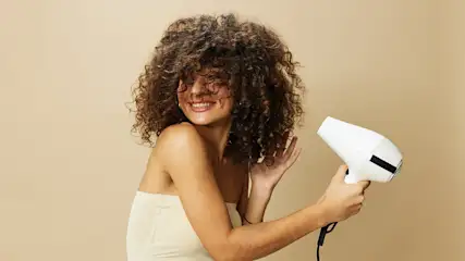 Frau im weißen Top, lächelnd, mit lockigem Haar. Sie hält einen weißen Föhn vor einem beigen Hintergrund.