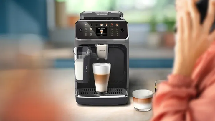 Kaffeemaschine mit Milchbehälter und Latte Macchiato im Glas. Person telefoniert im Hintergrund.