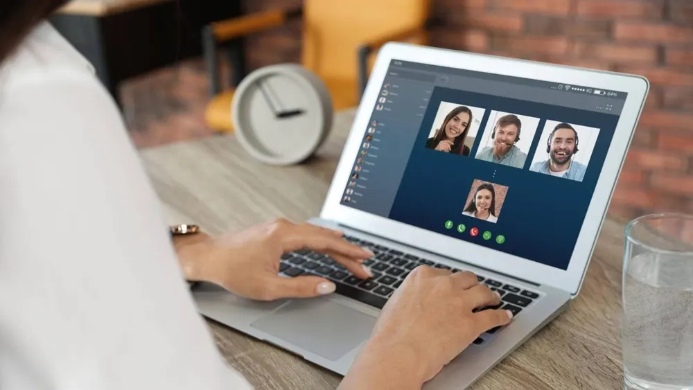 Eine Person tippt auf einem Laptop, der eine Videokonferenz mit vier Personen zeigt. Eine Uhr und ein Glas Wasser stehen auf dem Tisch.