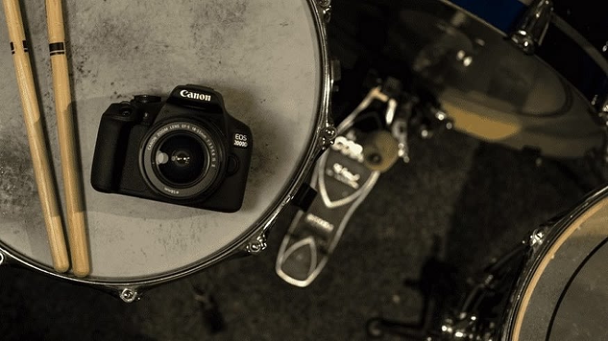 CANON EOS 2000D Kit Spiegelreflexkamera, 24,1 Megapixel, 18-55 mm Objektiv (EF-S, IS II), WLAN, Schwarz liegt auf einem Schlagzeug neben Drumsticks