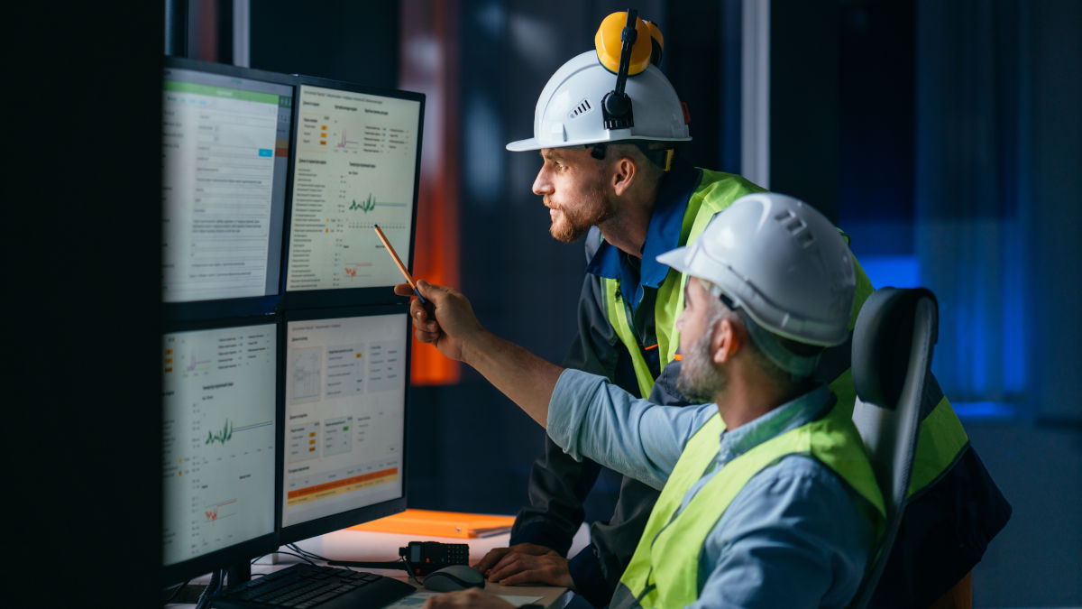 Zwei Bauarbeiter mit Helmen und Warnwesten betrachten Diagramme auf vier Monitoren und zeigen mit einem Bleistift darauf.