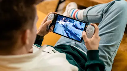 Eine Person sitzt und spielt ein Videospiel auf einem Tablet mit Controllern. Sie trägt Jeans und Socken.