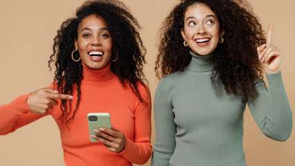 Weibliche Zwillinge: Eine Frau zeigt auf das Handy in ihrer Hand. Die andere Frau sieht aus, als hätte sie eine Idee.