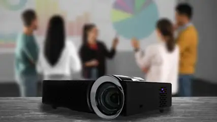 Schwarzer Projektor auf Tisch, im Hintergrund verschwommene Personen vor Diagramm auf Leinwand. Büroumgebung mit Präsentation.