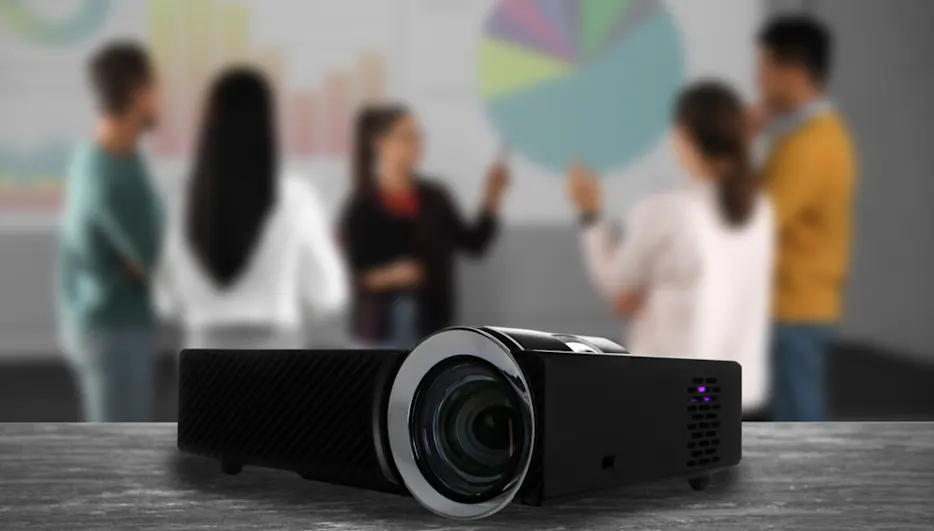 Schwarzer Projektor auf Tisch, im Hintergrund verschwommene Personen vor Diagramm auf Leinwand. Büroumgebung mit Präsentation.