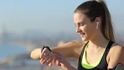 Frau im Sport-BH mit Ohrhörern, die auf eine Smartwatch schaut, mit Stadtbild im Hintergrund.