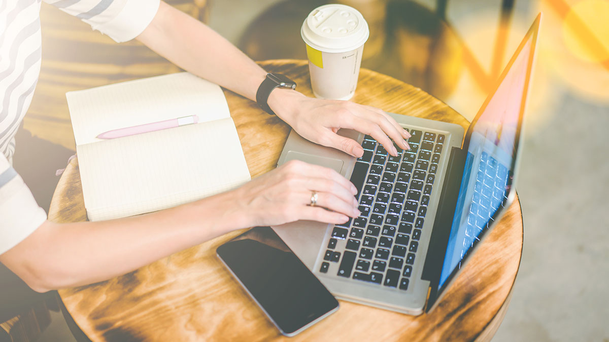 Eine Person tippt auf einem Laptop mit beleuchteter Tastatur auf einem Holztisch, daneben ein Notizbuch mit Stift, ein Smartphone und ein Kaffeebecher.