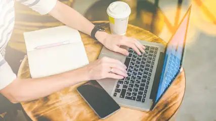 Eine Person tippt auf einem Laptop mit beleuchteter Tastatur auf einem Holztisch, daneben ein Notizbuch mit Stift, ein Smartphone und ein Kaffeebecher.