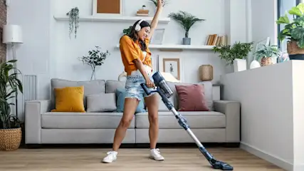 Frau mit Kopfhörern, die mit einem Staubsauger in einem Wohnzimmer mit Sofa und Pflanzen tanzt.