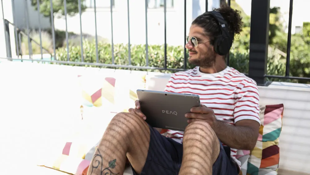 Ein Mann mit Kopfhörern und Sonnenbrille sitzt draussen und hält ein graues Tablet. Er trägt ein rot-weiss gestreiftes Hemd.