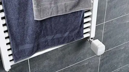 Ein weißer Handtuchwärmer hängt an einer gefliesten Wand. Ein blaues und ein graues Handtuch hängen darüber.