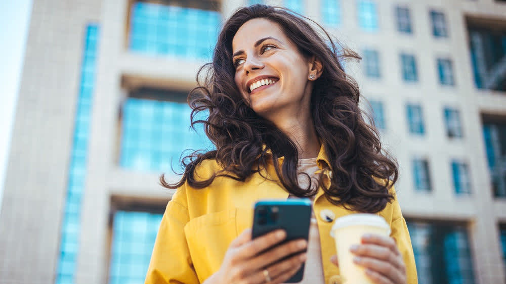 Eine lächelnde Frau hält einen Kaffee und ein Smartphone in den Händen.