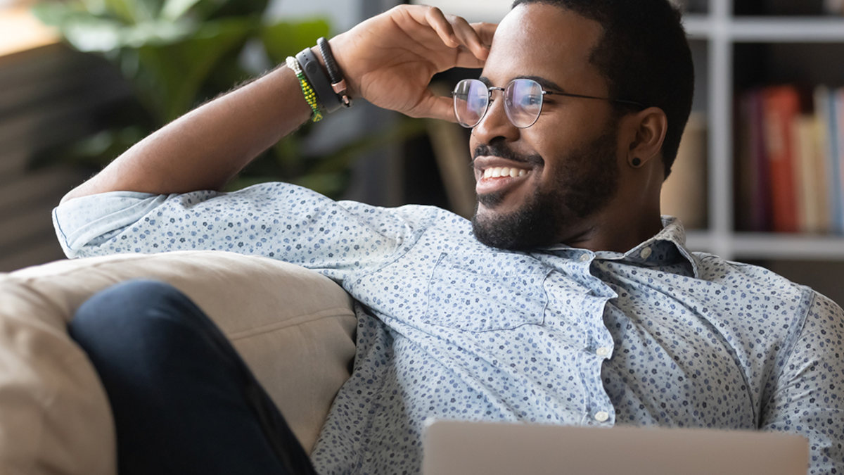 Ein Mann mit Brille lehnt lächelnd auf einem beigen Sofa; er trägt ein helles Hemd und dunkle Armbänder. Im Hintergrund stehen Bücher.