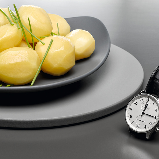 Gelbe Kartoffeln mit Schnittlauch auf einem grauen Teller, gestapelt auf einem weiteren Teller, neben einer silbernen Uhr.