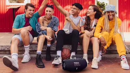 Fünf lächelnde Personen sitzen vor einer roten Wand. Ein Basketball und ein JBL-Lautsprecher stehen auf dem Boden.