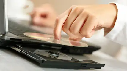 Eine Hand legt eine CD in ein geöffnetes CD-Laufwerk eines schwarzen Laptops. Der Laptop steht auf einem hellen Tisch.