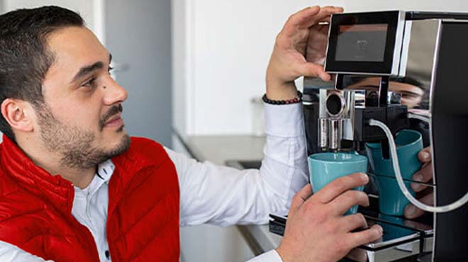 Ein Mann bedient eine Kaffeemaschine, hält eine blaue Tasse und drückt einen Knopf. Er trägt eine rote Weste und ein weißes Hemd.