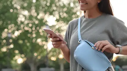Eine Person hält ein Smartphone und trägt eine blaue Gürteltasche über einem grauen Oberteil, Bäume im Hintergrund.