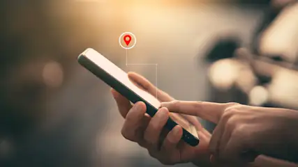 Eine Person hält ein Smartphone mit einem roten Standort-Pin auf dem Bildschirm. Der Hintergrund ist unscharf.