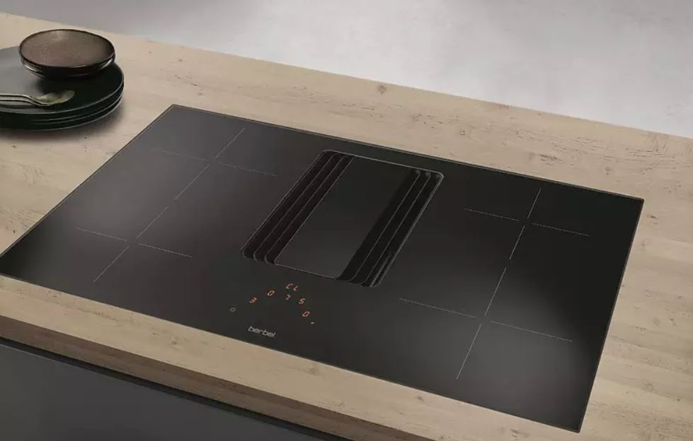 Schwarzes Kochfeld mit integriertem Dunstabzug auf einer hellen Arbeitsplatte. Tellerstapel und Löffel links oben.