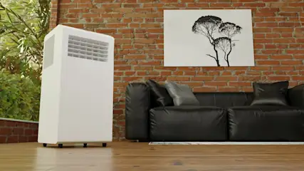 Ein weißes Klimagerät steht neben einer schwarzen Couch vor einer Ziegelwand mit einem Baumkunstdruck.