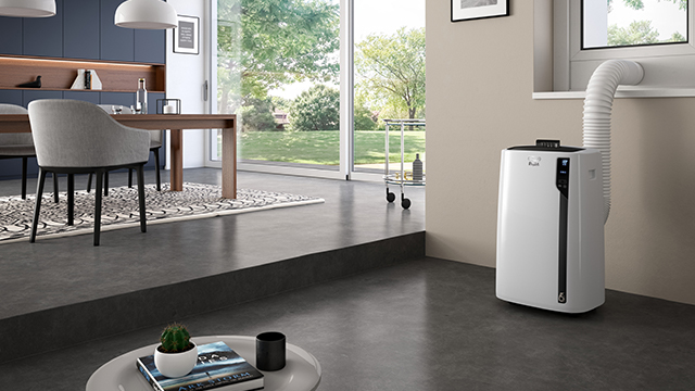 Ein Raum mit Esstisch, Stuhl und Klimaanlage am Fenster. Ein kleiner Tisch mit Pflanze und Büchern steht davor.