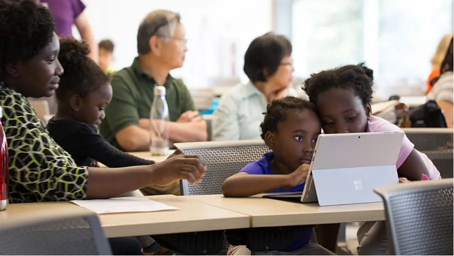 In einem Klassenzimmer sitzen mehrere Personen an Tischen, darunter Kinder, die ein Tablet benutzen.