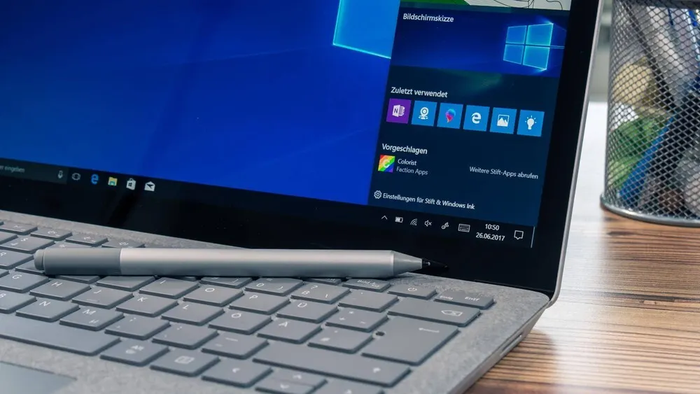 Ein grauer Stift liegt auf der Tastatur eines Laptops. Der Laptop zeigt den Windows-Startbildschirm. Ein Stiftehalter steht daneben.