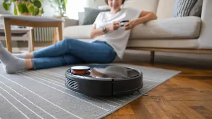 Eine Frau entspannt auf einem Sofa, während ein schwarzer Staubsaugerroboter auf einem gestreiften Teppich reinigt.