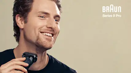 Ein Mann im schwarzen T-Shirt lächelt und hält einen Braun-Rasierer an seinen Hals vor einem beigen Hintergrund.